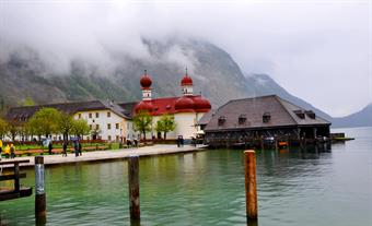 A red-roof church and several other building are situated in front of mountains. In the foreground there is green water of a lake, which surrounds the buildings.